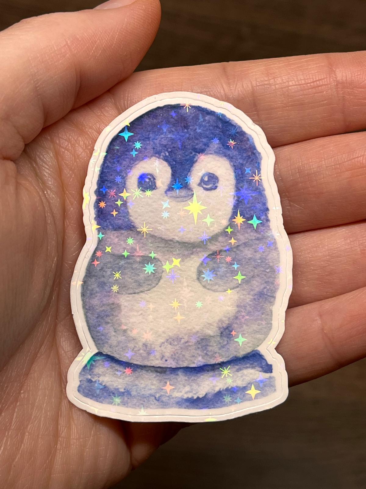Glitzernder Sticker "Kleiner Kaiserpinguin" in der Hand haltend